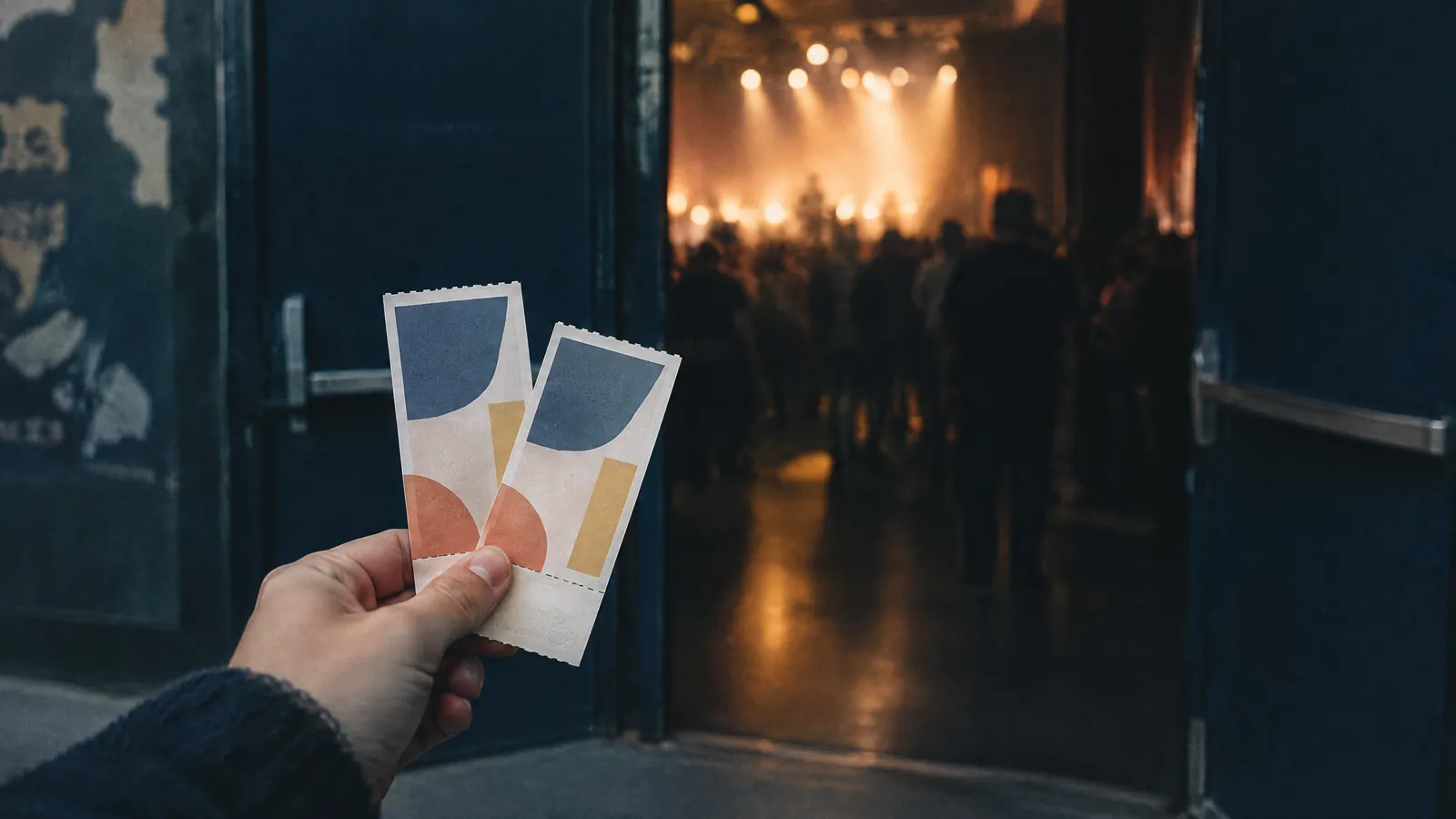 Hand holding two tickets at the entrance to a lit concert hall