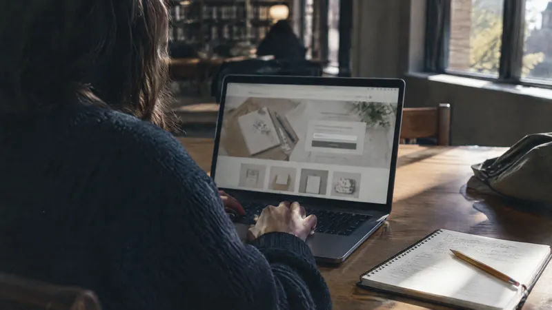 Person at a laptop in a library, notebook and pencil on the table