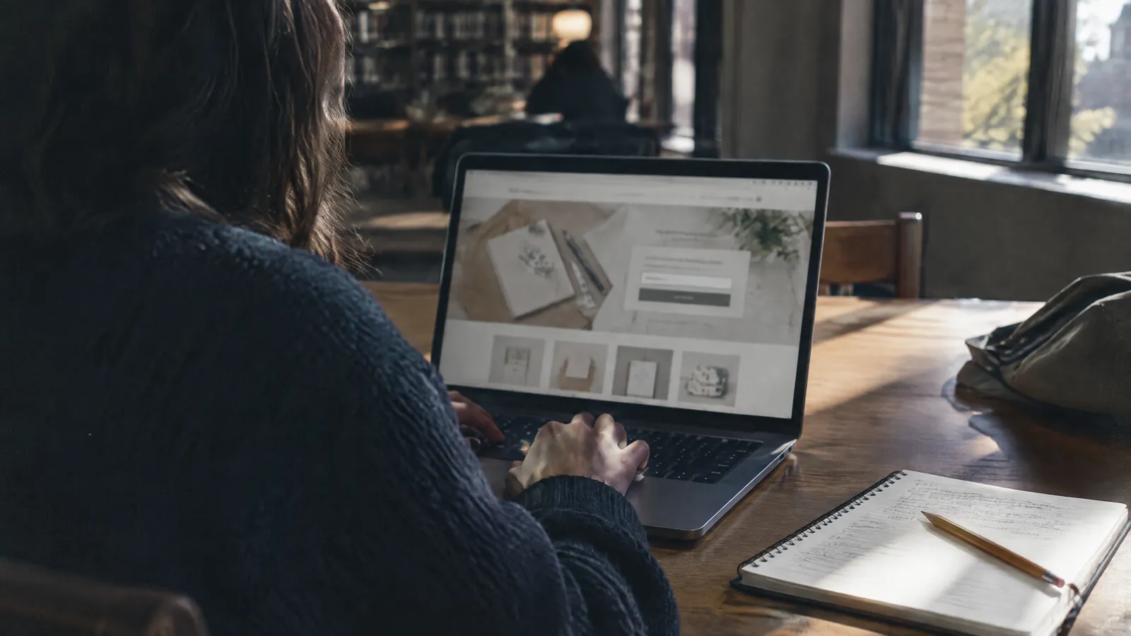 Person at a laptop in a library, notebook and pencil on the table
