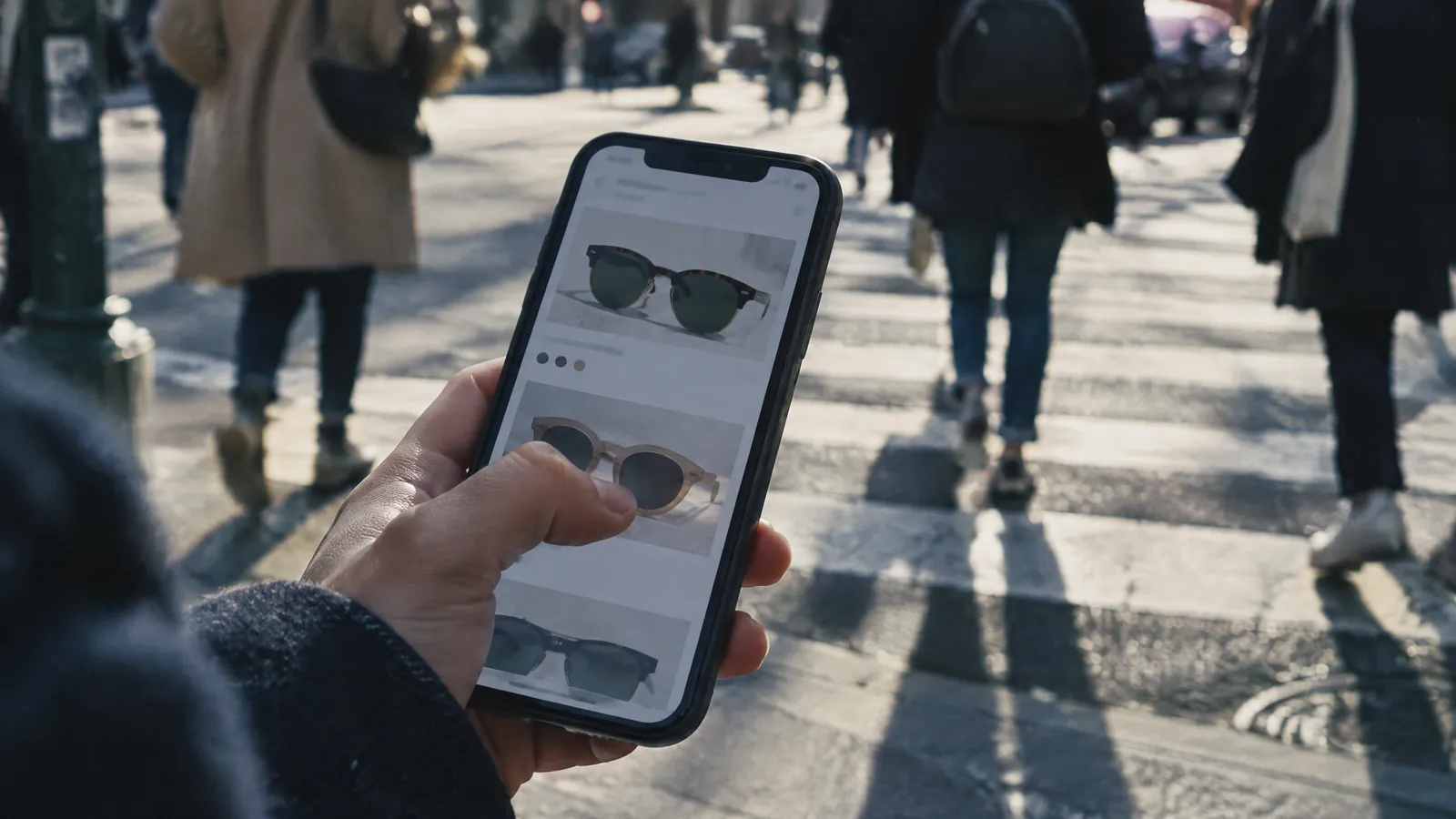 Hand holding a phone showing sunglasses while crossing a busy street