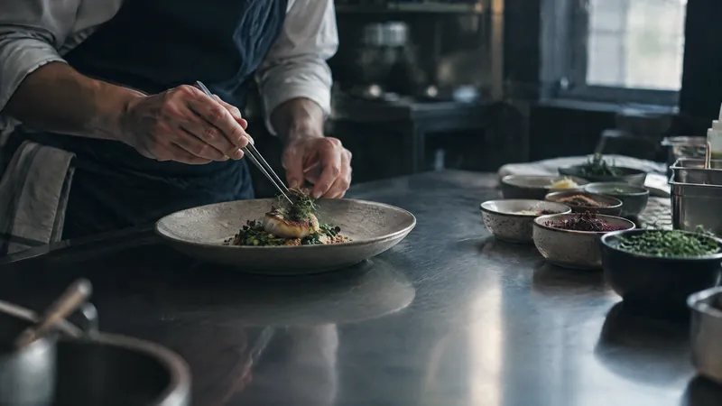 Chef plating a dish with tweezers in a kitchen lined with prep bowls