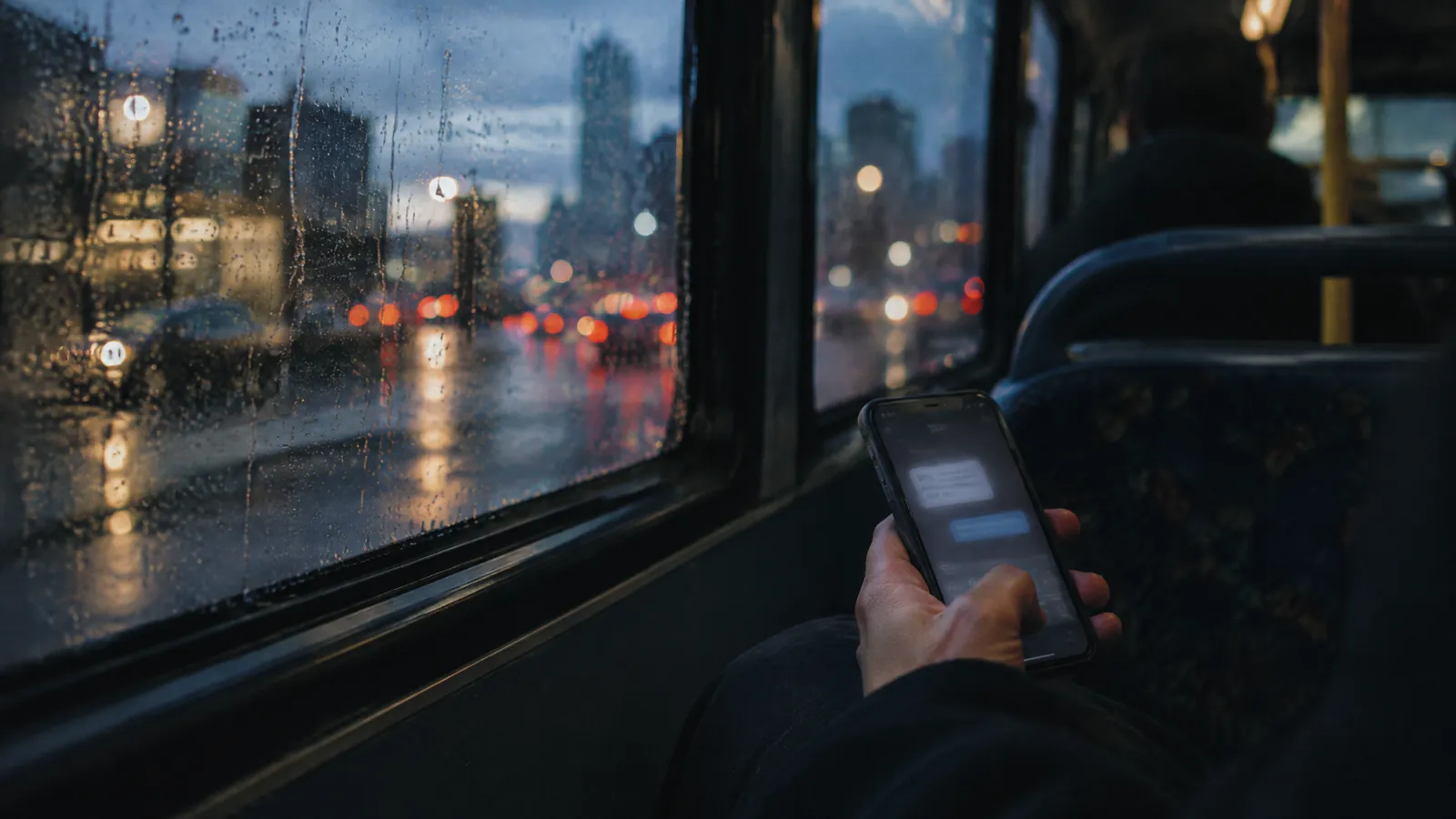 Hand holding a phone on a bus beside a rain-streaked window at night